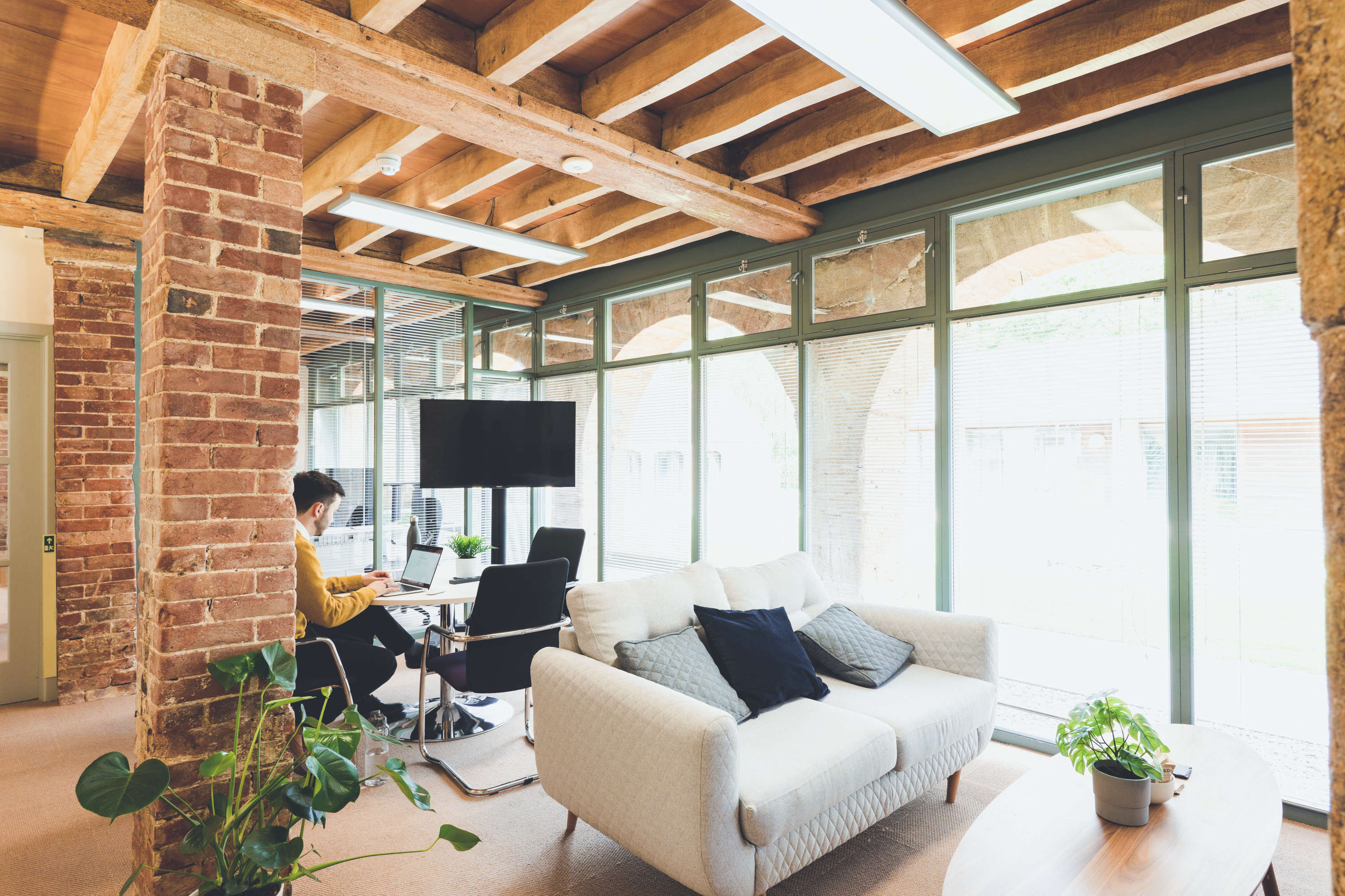 A rustic modern interior office space with fabric sofa, coffee table and a large glass wall. A project manager conducts a video call.